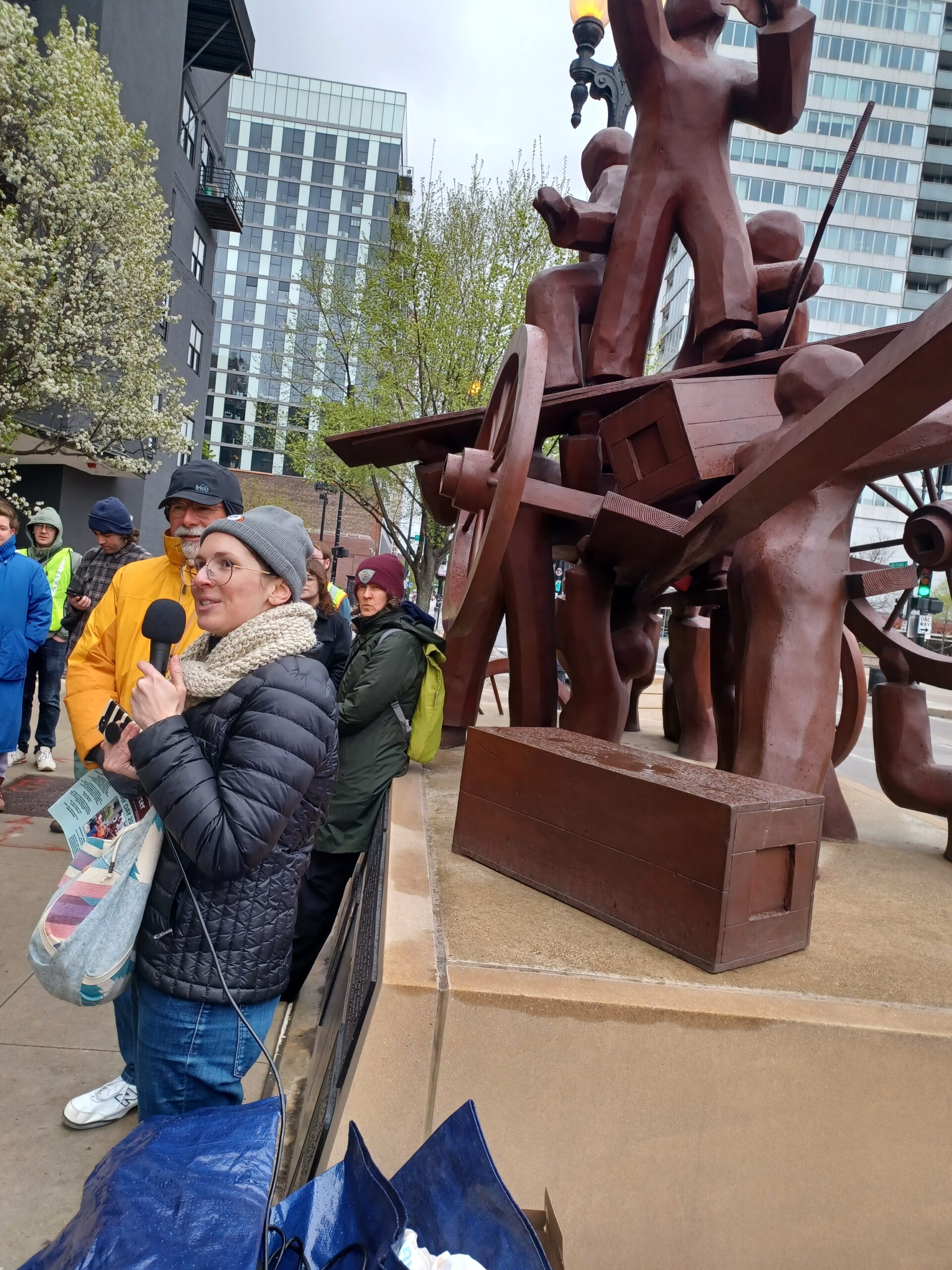 Une personne en tenue d'hiver tient un micro devant le mémorial Haymarket de Chicago, une statue brune représentant des personnes au rendu abstrait construisant ensemble un wagon.